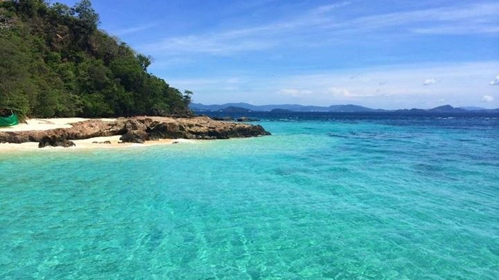 Tropical beach with clear turquoise water, rocky shoreline, and lush greenery under a blue sky.