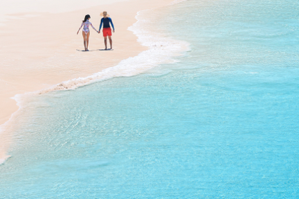 A couple walks along a sandy beach by clear blue water.