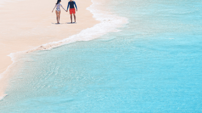 A couple walks along a sandy beach by clear blue water.