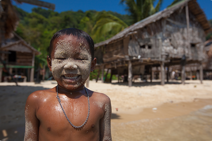 Child with sand on face smiling on a tropical beach near wooden huts.