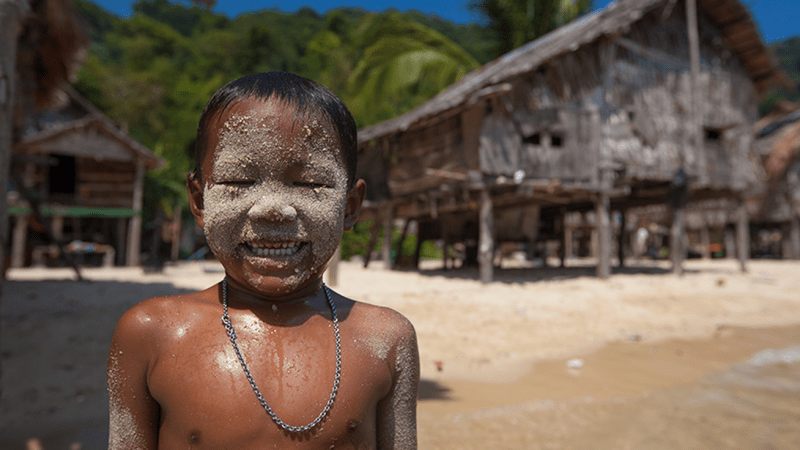 Child with sand on face smiling on a tropical beach near wooden huts.