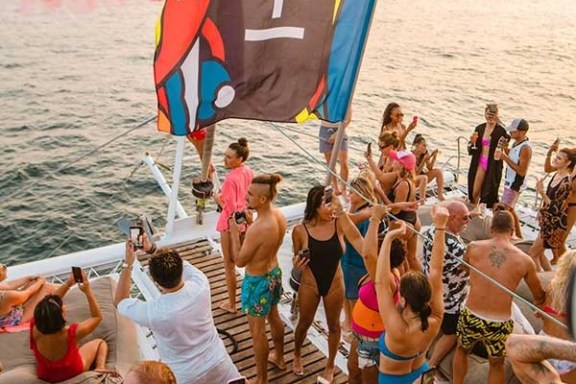 People partying on a boat deck with a colorful flag and sunset over the ocean.