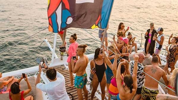 People partying on a boat deck with a colorful flag and sunset over the ocean.