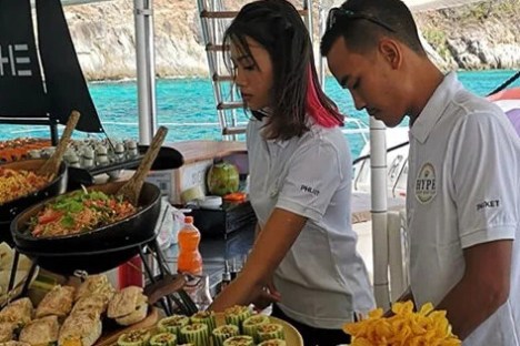 Two people serve food from a buffet on a boat with turquoise water in the background.
