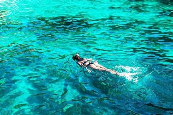 Person snorkeling in clear turquoise water near a rocky shore.