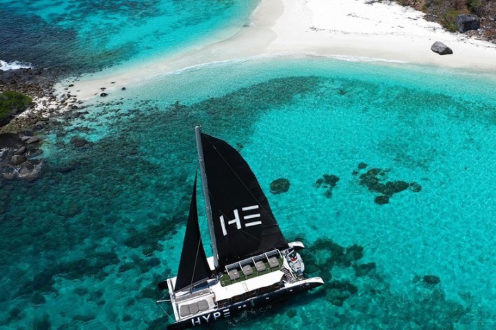 Aerial view of a catamaran sailing on clear turquoise water near a rocky, forested coastline with a sandy beach.
