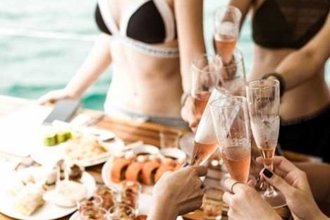 People in swimwear toasting with champagne on a yacht near a table of food.