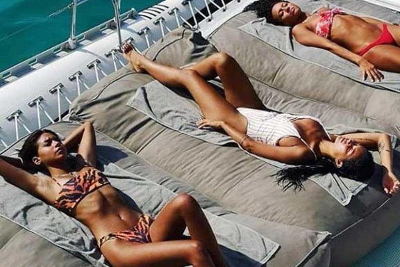 Three women sunbathing on a yacht, each wearing a swimsuit and lying on cushions.