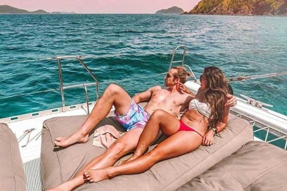 Couple lounging on a boat under a pink sky, with ocean and hills in the background.