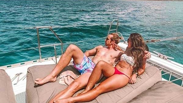 Couple lounging on a boat under a pink sky, with ocean and hills in the background.